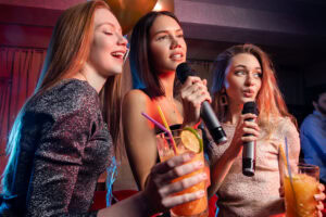 Three young women singing karaoke and holding microphones and cocktails, smiling and enjoying themselves at a party with festive lighting in the background.