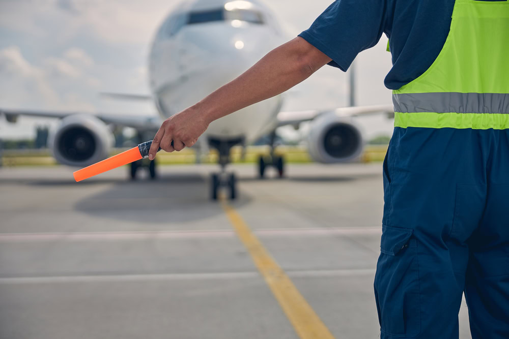 An airport ground crew member in a reflective vest signals with an orange wand to guide a commercial airplane as it taxis on the runway.