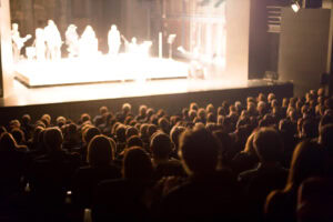 A blurred image of an audience seated in a dark theater, watching a brightly lit stage with several indistinct performers standing on it.