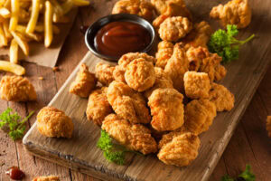 A wooden board topped with crispy fried chicken nuggets, fresh parsley, and a small bowl of barbecue sauce, with a side of French fries in the background.