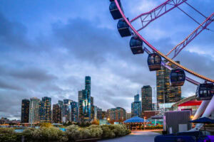 A large Ferris wheel dominates the right side of the image, with modern skyscrapers in the background under a cloudy evening sky in an urban waterfront setting.