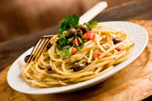 A white plate of spaghetti pasta garnished with black olives, chopped tomatoes, fresh parsley, and a gold fork resting on the side.