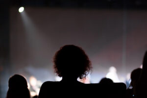 A silhouette of a person with curly hair seated in a dark theater, facing a blurred, brightly lit stage with other audience members visible in the foreground.
