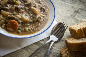 A bowl of hearty stew with vegetables and meat sits on a wooden table beside a napkin, a fork, a spoon, and three slices of bread.