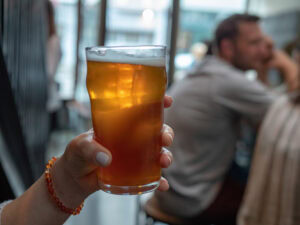 A hand holding a pint glass of amber beer in a dimly lit indoor setting, with people sitting and chatting in the blurred background.
