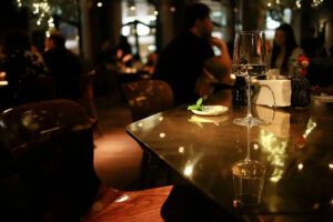 A dimly lit restaurant with a glass of white wine on a reflective table, surrounded by blurred diners. Warm lighting and string lights create a cozy, intimate atmosphere.