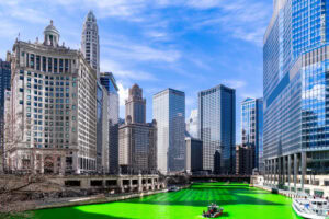 Chicago skyline with tall buildings along the river, which has been dyed bright green for St. Patrick’s Day; a small boat is visible on the water under a blue sky.