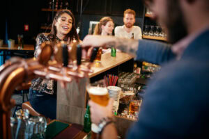 A bartender pours beer from a tap for a smiling woman sitting at the bar, while other people socialize in the background in a lively, modern bar setting.