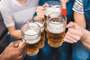 Four people clinking large glass mugs of beer together in a toast, viewed from above. The image shows only their hands and part of their arms, with light foam visible on the beers.