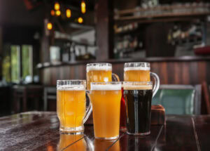 Five glasses of beer, each with different colors and levels of foam, are arranged on a wooden table in a dimly lit bar with hanging lights and shelves in the background.