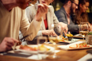 A group of people sit at a table in a restaurant, enjoying food and drinks. The focus is on their hands and plates, with dishes and glasses of wine visible. Faces are partially blurred, creating a lively dining atmosphere.