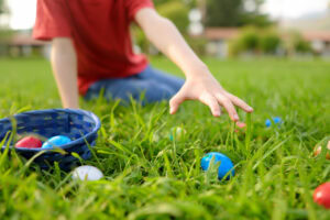 A child in a red shirt kneels on grass, reaching for colorful eggs scattered near a blue basket during an outdoor egg hunt.