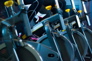 Close-up of several stationary exercise bikes in a gym, with people pedaling, wearing athletic clothing and sneakers, focused on the legs and bike equipment.