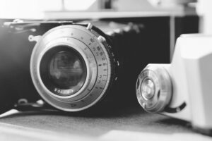 Close-up, black-and-white image of a vintage camera lens and a light meter placed on a surface, highlighting the detailed dials and metallic texture.