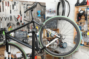 A bicycle is mounted on a repair stand in a workshop filled with various tools, bike wheels, and parts organized on shelves and pegboards.