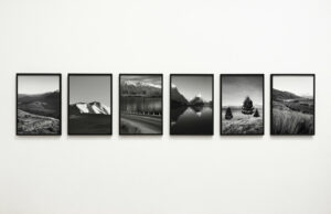 Six black-and-white framed photographs of landscapes, including mountains, water, trees, and winding roads, are displayed in a horizontal row on a white gallery wall.