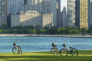 Three bicycles are parked on a grassy area by the water, with a city skyline of tall buildings and trees in the background under clear daylight.