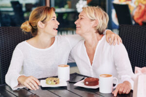 Two women smiling and embracing each other while sitting at a table with coffee and cakes, enjoying a friendly moment outdoors.