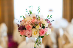 A close-up of a floral arrangement with pink and white roses, small purple flowers, and greenery in a clear glass vase, set against a softly blurred indoor background.