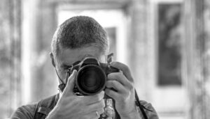 A person with short hair and glasses is holding a camera up to their face, taking a photo. The image is in black and white, with a blurred background and framed artwork behind them.