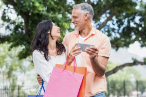 A smiling couple stands outdoors holding colorful shopping bags and a tablet, looking at each other happily with trees and greenery in the background.