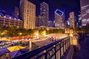 A vibrant cityscape at night with tall, illuminated skyscrapers and a lively outdoor market below, featuring colorful tents, string lights, and trees, viewed from behind a decorative railing.