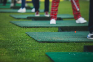Several people stand on individual green golf mats on a grassy field, practicing their swings. Only their lower legs and feet are visible, with one person wearing red plaid pants.