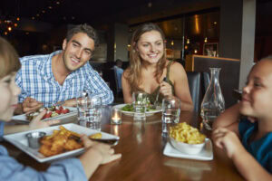 A smiling family of four sits at a restaurant table, enjoying a meal together. Two adults and two children are eating and talking, with plates of food, glasses of water, and a candle on the wooden table.