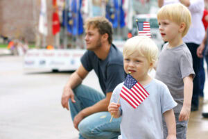 Two young boys and a man watch a parade. The boys hold small American flags, and one has a flag tucked behind his ear. The scene appears festive with blurred flags and people in the background.
