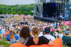 People sitting on colorful bean bags watch a large outdoor concert. A crowd gathers near the stage set up with lights and equipment, surrounded by greenery and trees in the background.