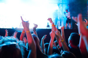 A crowd of people with raised hands and wristbands enjoy a lively concert, illuminated by bright stage lights and blue tones, with a stage and photographer visible in the background.
