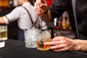 A bartender stirs a cocktail with ice in a glass, holding a mixing spoon and a jigger, while another glass filled with ice sits nearby on the bar countertop. Bottles are visible blurred in the background.