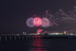 Fireworks burst in red and blue above a lit-up waterfront at night, with a ferris wheel and buildings in the distance and their reflections shimmering on the water.