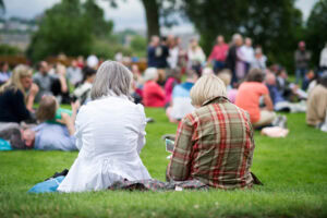 Two people sit on grass with their backs to the camera at an outdoor event. A crowd of people is gathered in the background, and trees frame the scene, suggesting a park or garden setting.