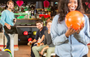 Four friends are at a bowling alley. Three are sitting with drinks, smiling at the camera, while one person in the foreground holds an orange bowling ball, out of focus and smiling. The background shows bowling balls and a bar.