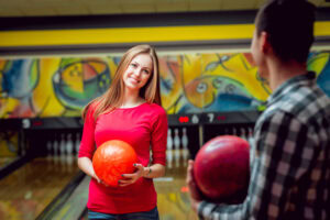 A young woman in a red shirt holds an orange bowling ball and smiles at a man holding a red ball in a bowling alley with colorful wall art in the background.