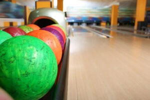 A close-up of colorful bowling balls in a rack, with a bowling lane and pins visible in the blurred background of a bowling alley.