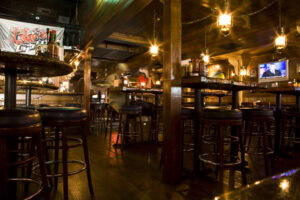 Dimly lit bar interior with high wooden tables and stools, hanging lantern lights, bottles on tables, and a TV mounted on the wall showing a news program. The atmosphere is warm and rustic.