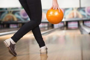 A person wearing bowling shoes is about to release an orange bowling ball down a bowling lane, with pins visible in the distance. Only the lower body and hand holding the ball are shown.