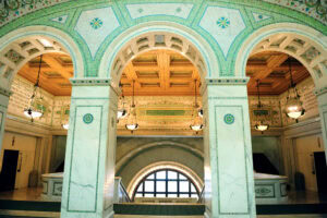 Ornate interior with three marble arches, intricate green and gold mosaic tiles, a coffered wooden ceiling, and vintage hanging lights above a staircase and large arched window.
