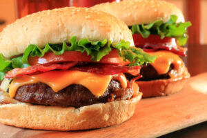 Two cheeseburgers on a wooden board, topped with lettuce, tomato, bacon, melted cheese, and nestled in sesame seed buns. The burgers appear fresh and appetizing.