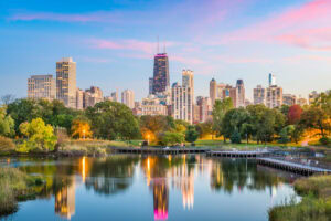Chicago skyline at sunset with skyscrapers, trees, and a park reflected in a calm pond under a colorful sky.