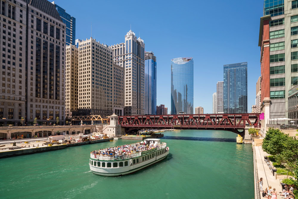 A boat carrying passengers cruises along a green river through downtown Chicago, surrounded by tall skyscrapers and a red iron bridge under a clear blue sky.