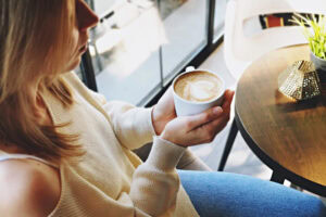 A woman in a cream sweater and jeans sits by a window, holding a cappuccino with latte art. A small table with a plant and a decorative object is beside her. Sunlight streams in through the window.