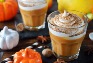 Two glasses of pumpkin spice smoothie topped with whipped cream and sprinkled spices, surrounded by mini pumpkins, a striped straw, and fall-themed decorations on a wooden table.