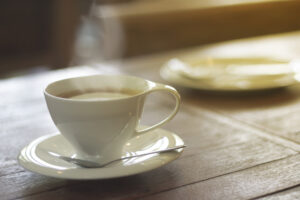 A white cup filled with coffee sits on a matching saucer with a spoon, placed on a wooden table. In the background, there is a blurred plate. Warm light shines from the side.