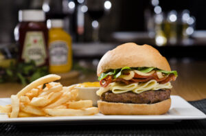 A juicy hamburger with lettuce, tomato, cheese, and bacon on a bun, served with a side of French fries and a small cup of sauce on a white plate; bottles of ketchup and mustard are blurred in the background.