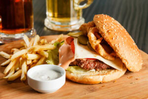 A cheeseburger with lettuce, tomato, pickles, onion rings, and sauce sits on a wooden board next to a pile of French fries and a small cup of dipping sauce, with a glass of beer in the background.
