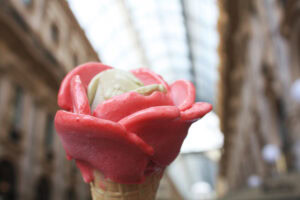 A close-up of an ice cream cone shaped like a pink rose, with a scoop of green ice cream in the center, held indoors under a glass-covered arcade with blurred architecture in the background.