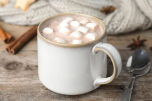 A white mug filled with hot chocolate topped with mini marshmallows sits on a wooden table near a metal spoon, cinnamon sticks, star anise, and a knitted gray blanket in the background.